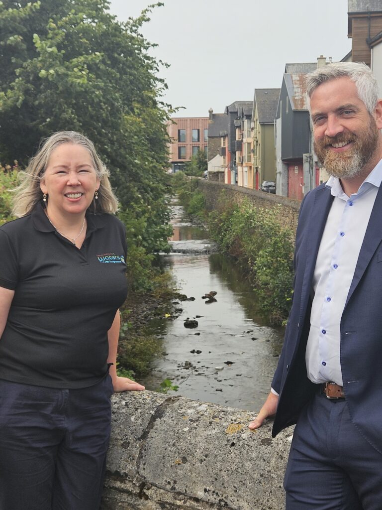 Two people standing near a small river or stream with buildings and trees in the background. 